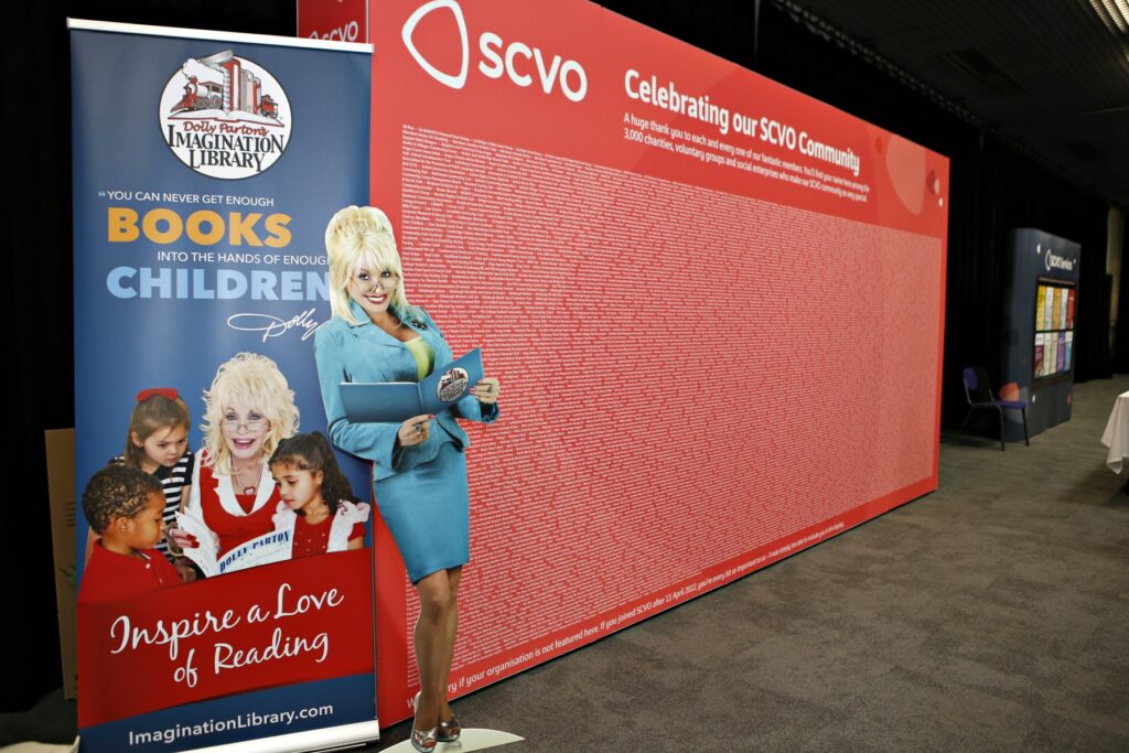 Blue pop up banner that reads: Dolly's Imagination Library. A cardboard cut out of country singer and philanthropist Dolly Parton leans against the pop up banner next to a large red canvas stand that says SCVO Celebrating our SCVO Community and lists 3000 SCVO community members on the stand in white writing