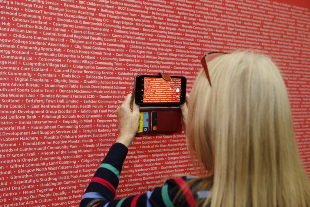 Person with blonde hair is seen from behind taking a photo of white writing on red canvas. The photo is seen on their phone screen