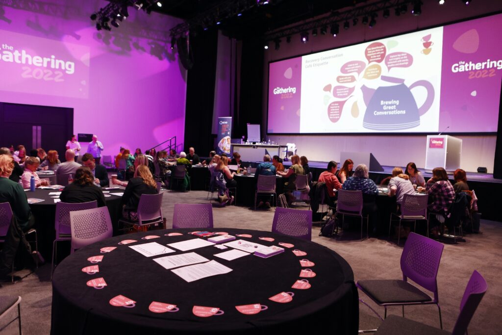 Large room with round tables and people seated at the tables, taking part in a conference. Large screen on the wall with a teapot image and the words "brewing great conversations" and "the Gathering 2022"