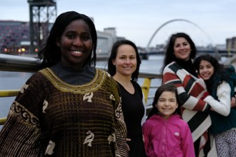 A group of 3 women and 2 children look directly to camera, smiling. They are standing on the banks of the Clyde in Glasgow
