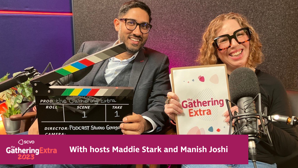 Manish and Maddie are sitting together , smiling n a podcast studio. Manish holding a clapperboard and Maddie is holding a Gathering Extra clipboard