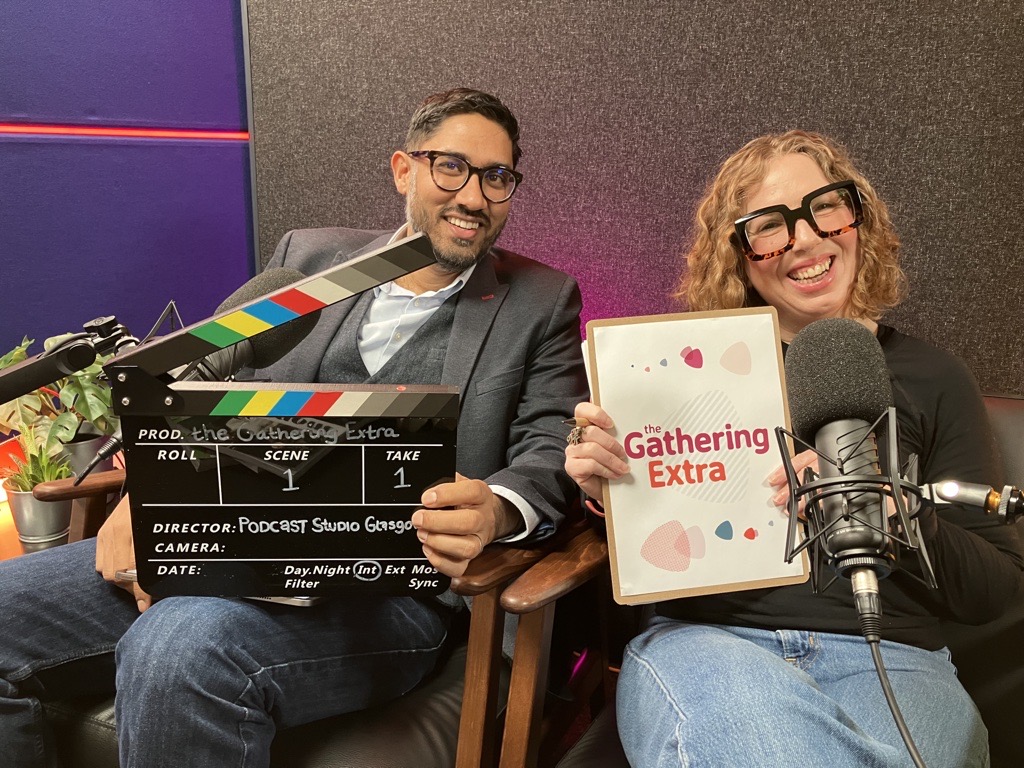 Manish and Maddie are sitting together , smiling n a podcast studio. Manish holding a clapperboard and Maddie is holding a Gathering Extra clipboard