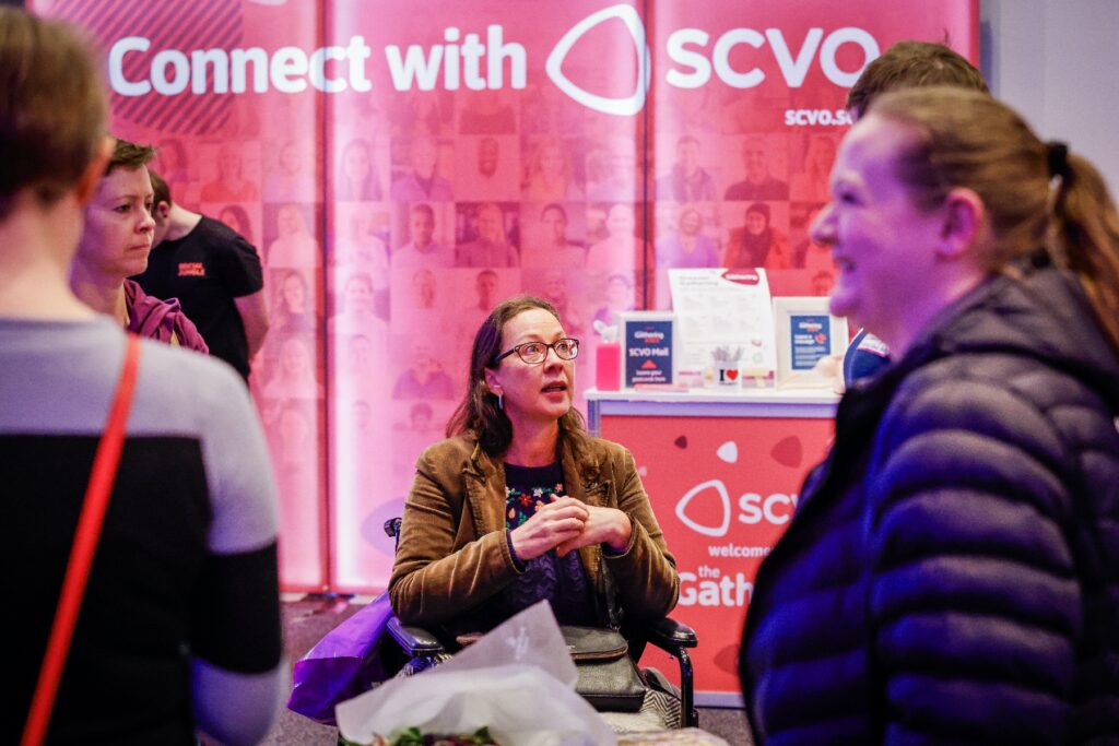 Delegates at a conference have conversations and smile in front of a graphic that reads "Connect with SCVO"