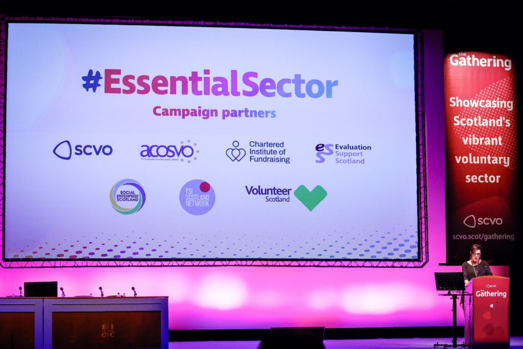 Woman at a lectern on a stage speaking. A screen behind her reads "#EssentialSector"