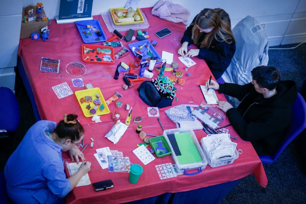 A group of people sit at a table with a red table cloth and arts and crafts materials, taking part in craft activities