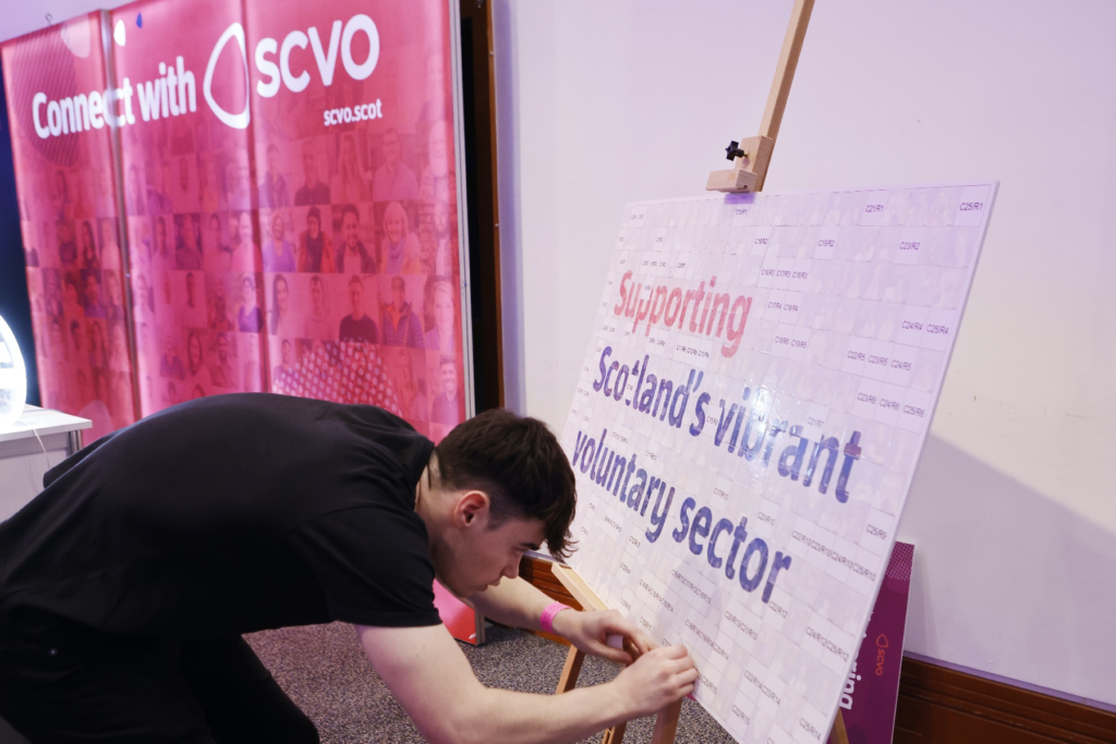 Light-up wall with "Connect with SCVO" written across it. A man in a black tshirt is attaching a sticker to a board with a mosaic of other stickers that spells out the words "Supporting Scotland's vibrant voluntary sector"