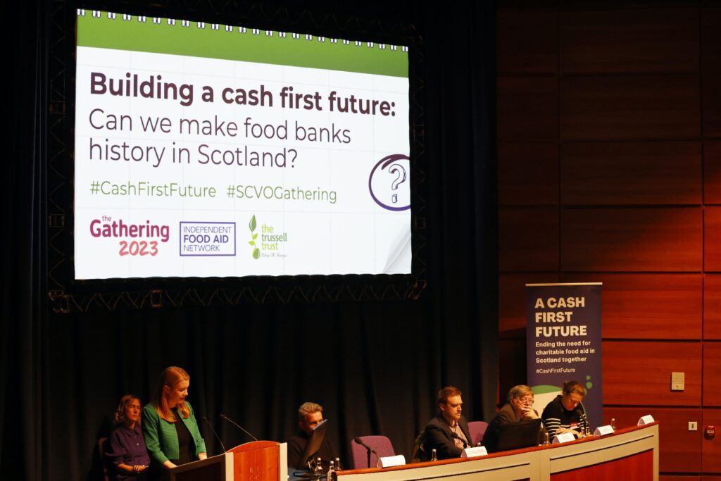 A woman in a green jacket (Shirley-Anne Somerville MSP, Cabinet Secretary for Social Justice) stands at a lectern delivering a speech. To her left is a panel of people sat at a table and behind her is a large screen that reads "Building a cash first future: can we make food banks history in Scotland?"