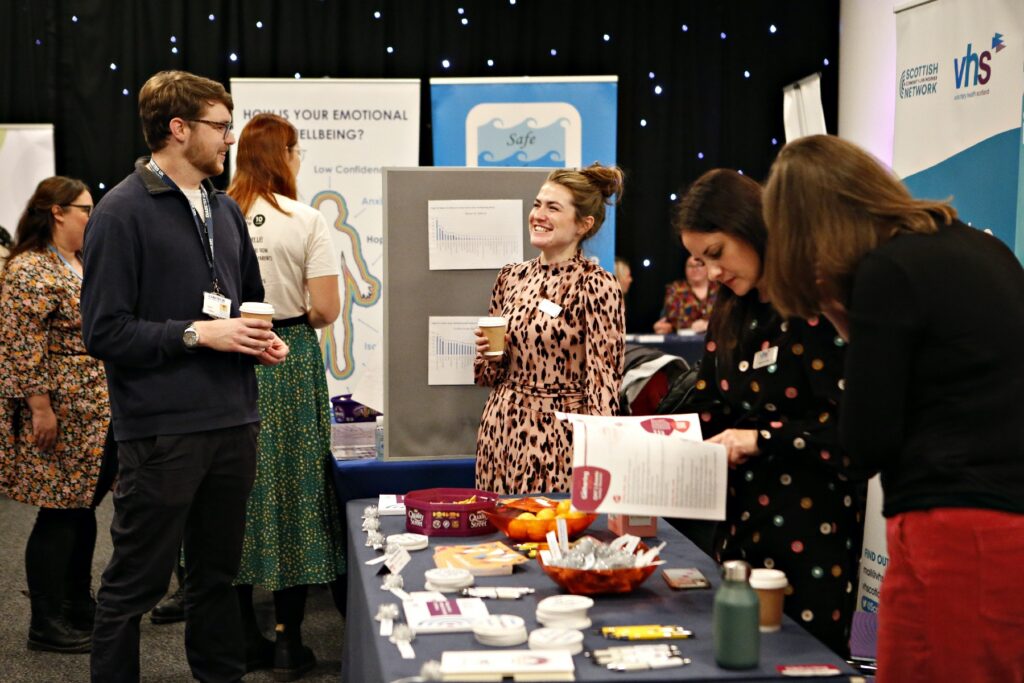 Two people holding coffee cups smile and chat at an exhibition table covered with a blue table cloth and promotional merchandise