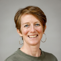 A headshot photo of Beth Mukushi - a woman with short, light brown hair and wearing silver hoop earrings smiles