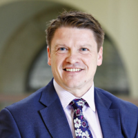 Headshot photo of Kenneth Pinkerton- a man with short brown hair, wearing a light pink button down collared shirt with a flowery tie and blue suit jacket smiling