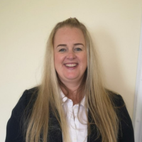 Headshot photo of Lucinda Godfrey- a woman with long blonde in a dark suit jacket, white blouse with long blonde hair smiling