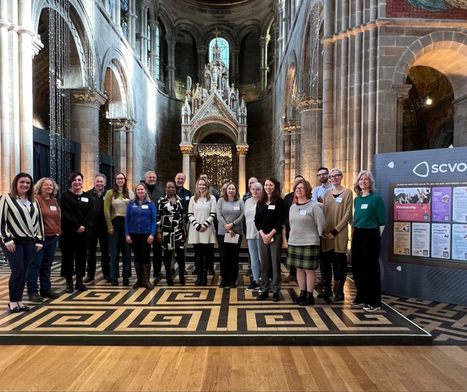 A group of about 20 people stand smiling at the camera next to a banner stand that reads "SCVO services!. THe group is standing in Mansfield Traquair Centre, an ornate former church now used as an office and events space