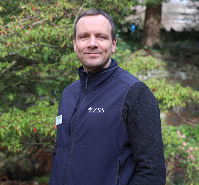 A man with brown hair smiles at the camera. He is wearing a blue gilet emblazoned with RZSS (Royal Zoological Society of Scotland). He is standing in front of some planting and trees