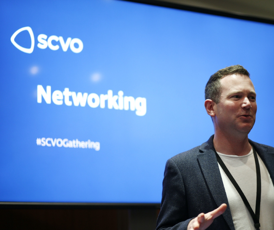 A man with brown hair waeing a white tshirt and blue blazer with a lanyard round his neck addresses a room. He is standing in front of a large blue screen. On the screen is a white 'SCVO' logo and the words 'Networking #SCVOGathering"