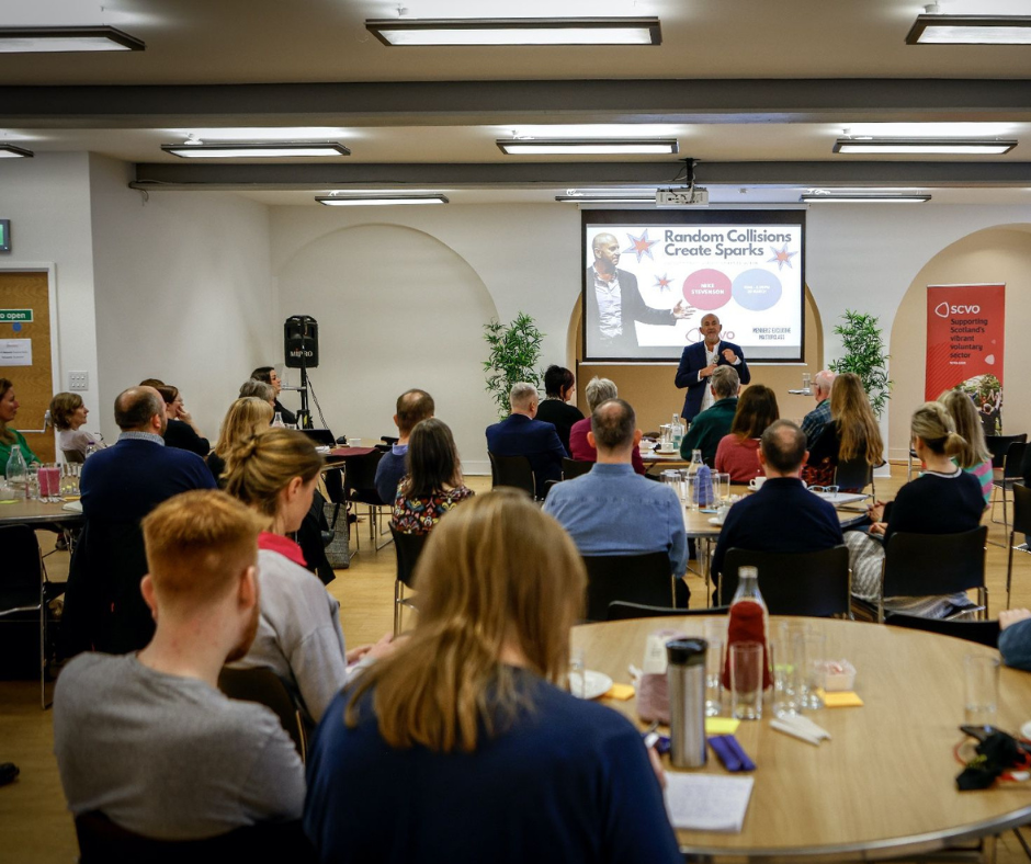 A man stands in front of a screen that has text that reads "Random collisions create sparks" written on it. The man is addressing a room full of people who are listening while they face him. They are seated a round tables