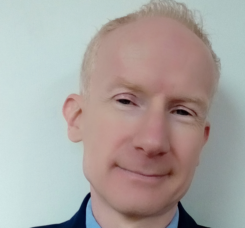 A headshot of a  man with light, closely cut hair smiles at the camera. He is wearing a shirt and a suit - only the collar can be seen