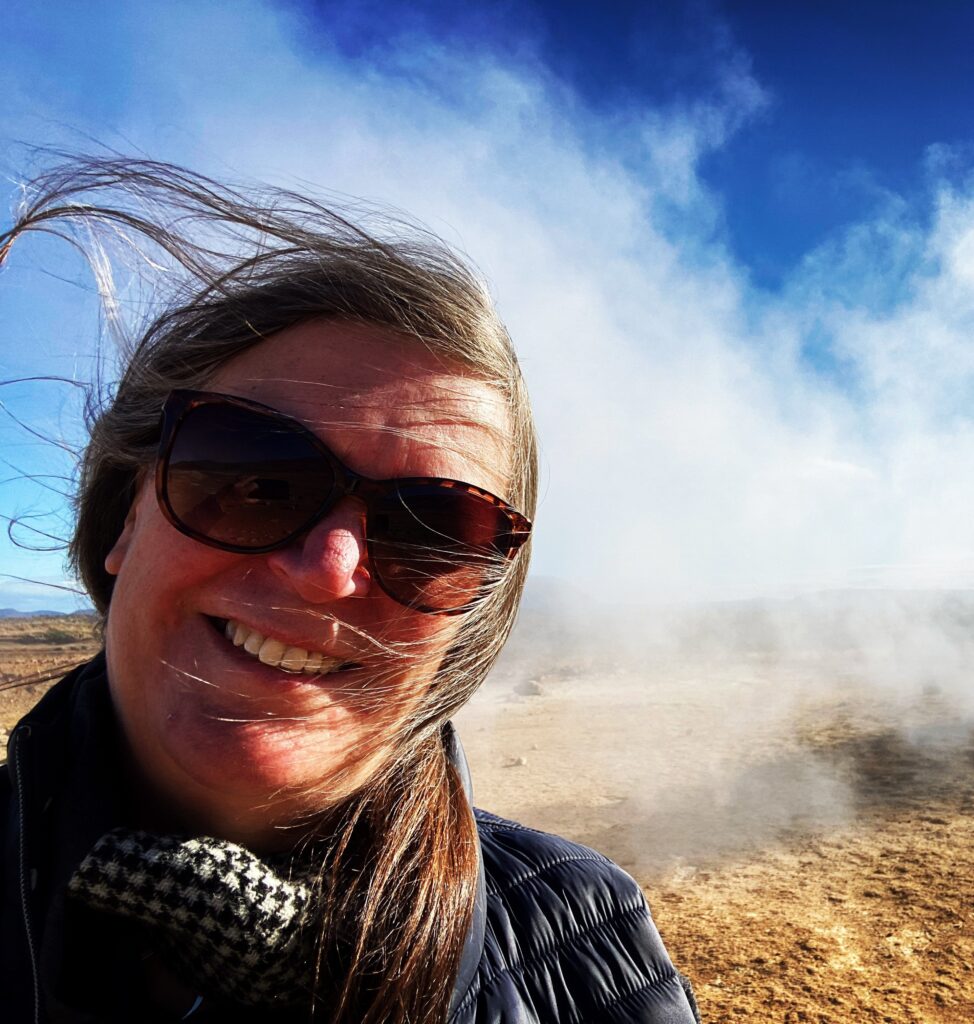Headshot of Carron Tobin, a white woman with brown hair wearing sunglasses and standing next to a geyser.