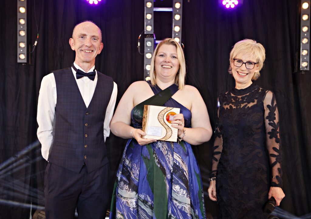 Three people standing on stage at the Scottish Charity Awards 2025. The woman in the centre, wearing a blue and green evening gown, is holding an award for Campaign of the Year and smiling. She is flanked by a man in a bow tie and vest on the left, and a woman in a black lace dress with glasses on the right. Stage lights and a black curtain backdrop are visible behind them