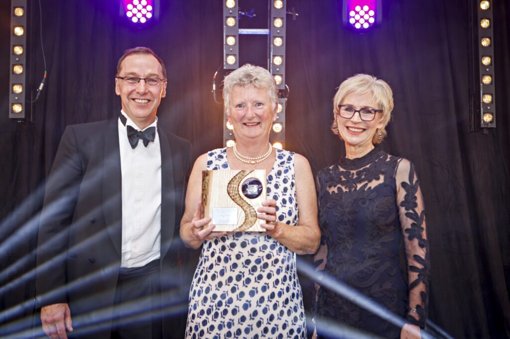 Three people on stage at the Scottish Charity Awards 2025. A woman in the centre, wearing a sleeveless white dress with blue floral patterns and a pearl necklace, is smiling and holding a wooden award plaque for the volunteer of the year award. She is flanked by a man in a tuxedo with a bow tie on the left and a woman in a dark, lacy evening dress and glasses on the right. Bright stage lights and a black curtain backdrop are visible behind them