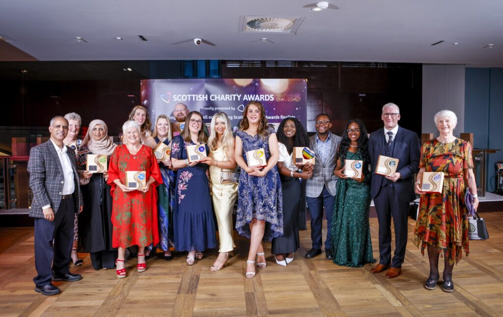Group photo of the winners at the Scottish Charity Awards 2025, posing indoors in formal attire. Each person is holding a unique wooden award plaque. A banner in the background reads 'Scottish Charity Awards 2025.' The group is diverse in age, gender, and ethnicity, celebrating their achievements together