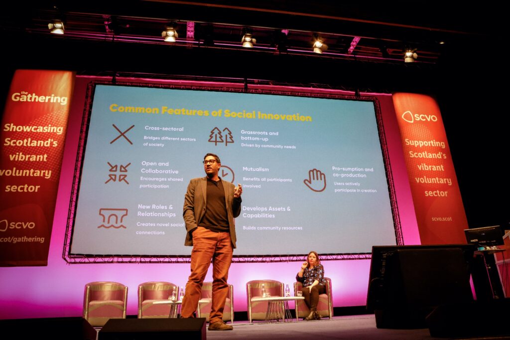 Manish Joshi presents on stage at the Gathering in front of a large screen with a presentation